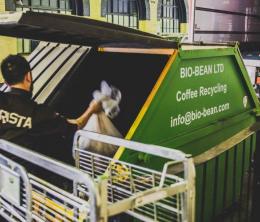 Man lifting a bag of coffee grounds into a recycling skip in a warehouse.