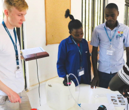 Tom Stakes, Blue Tap founder and Chief Technical Officer, holding a notepad and looking at a chlorine doser being tested in Uganda, watched by three members of the local water treatment project team, one female and two male. 