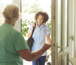 Elderly woman in light green top, with back to the camera, opening the door to community nurse, carrying black shoulder bag, arriving on a social care visit