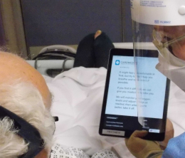 View from behind elderly patient's head of a nurse in full personal protection holding up a tablet with text explaining how oxygen treatment might make him feel. 