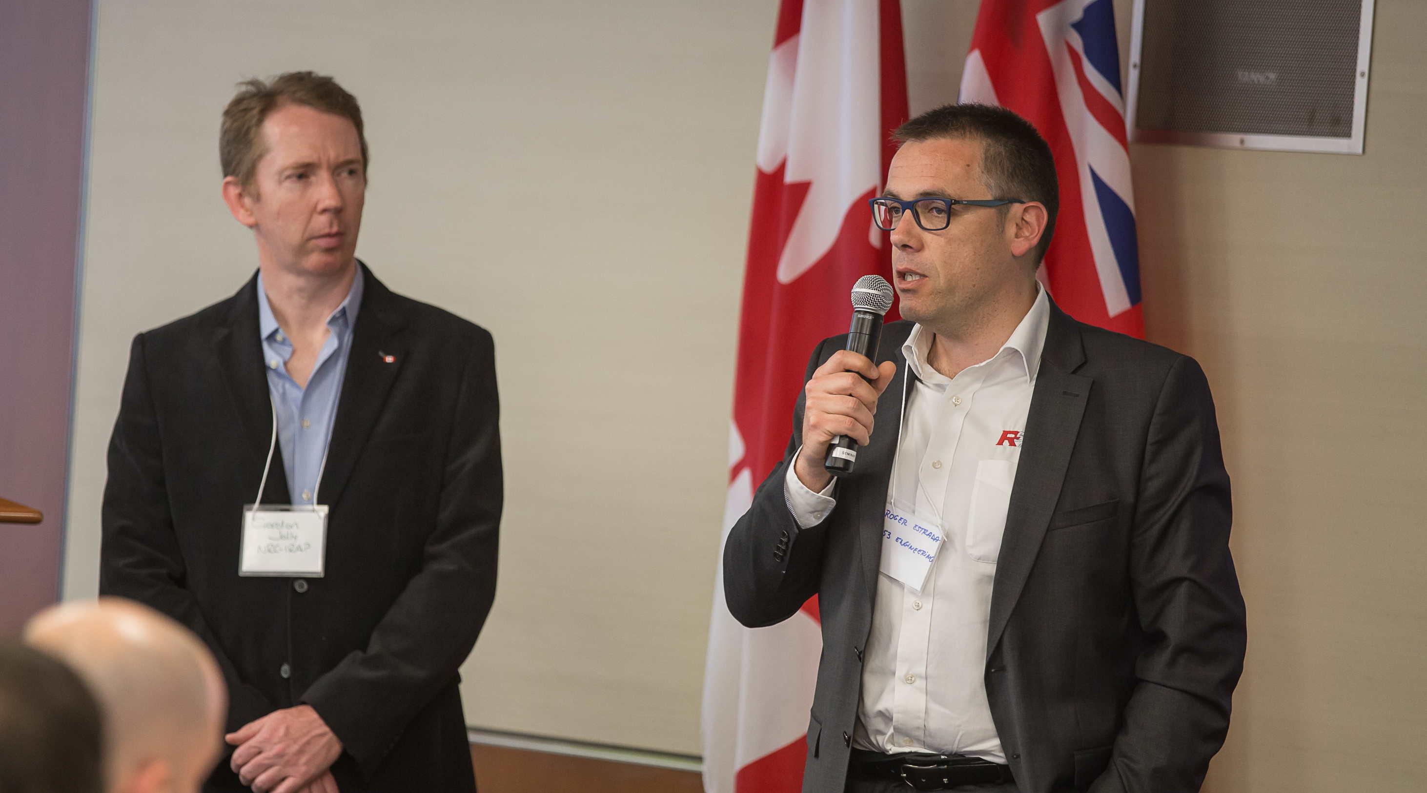 Roger Estrada, CEO of R53 Engineering, using a microphone to address an event during the GBIP visit to Canada. Looking on (left) is Gordon Jolly, ‎Industrial Technology Advisor - ‎National Research Council Canada. UK and Canadian flags on the wall behind.