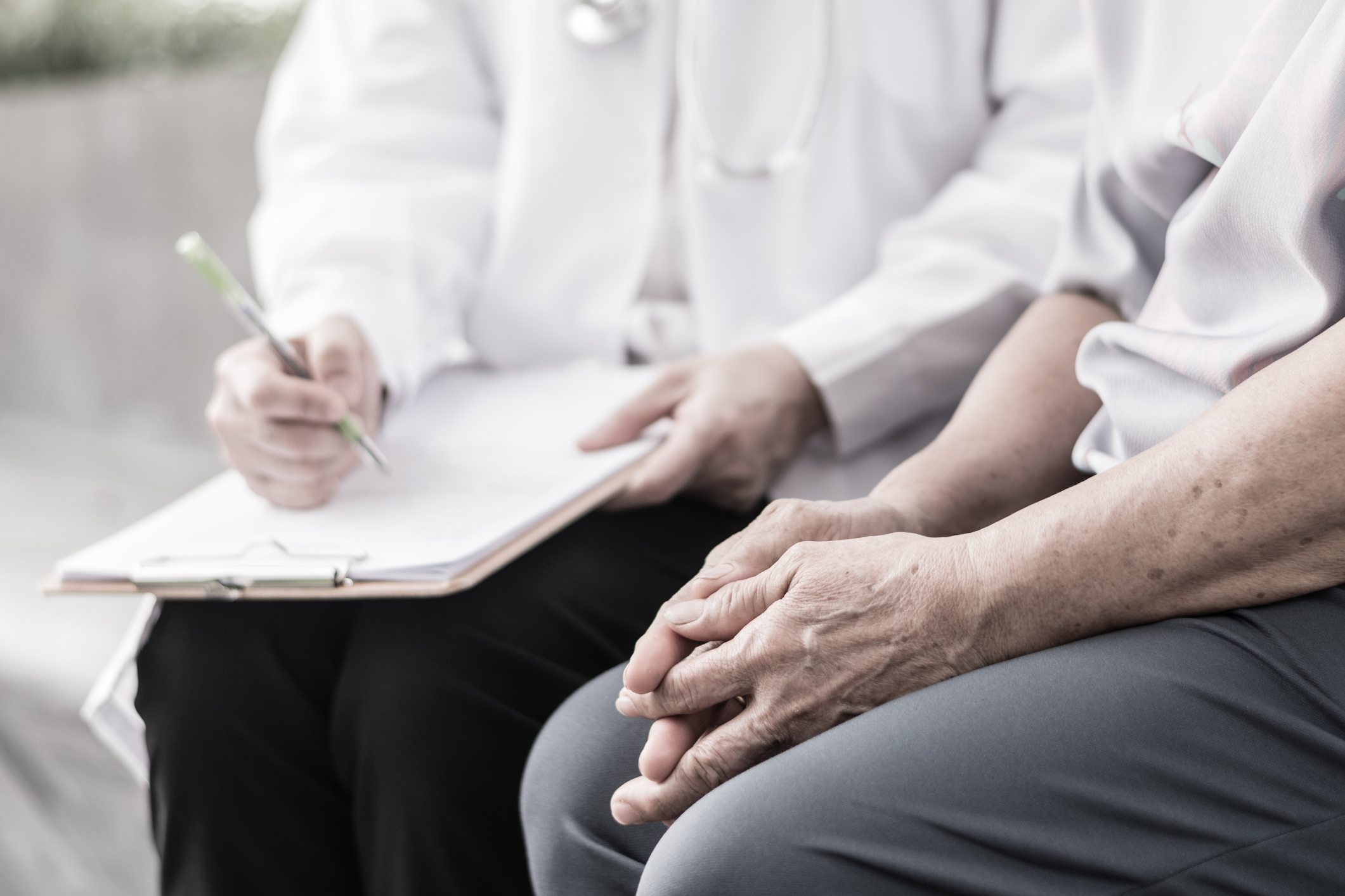 Close-up of elderly patient's hands clasped resting on his legs, with clinician in the background taking notes on a clipboard.