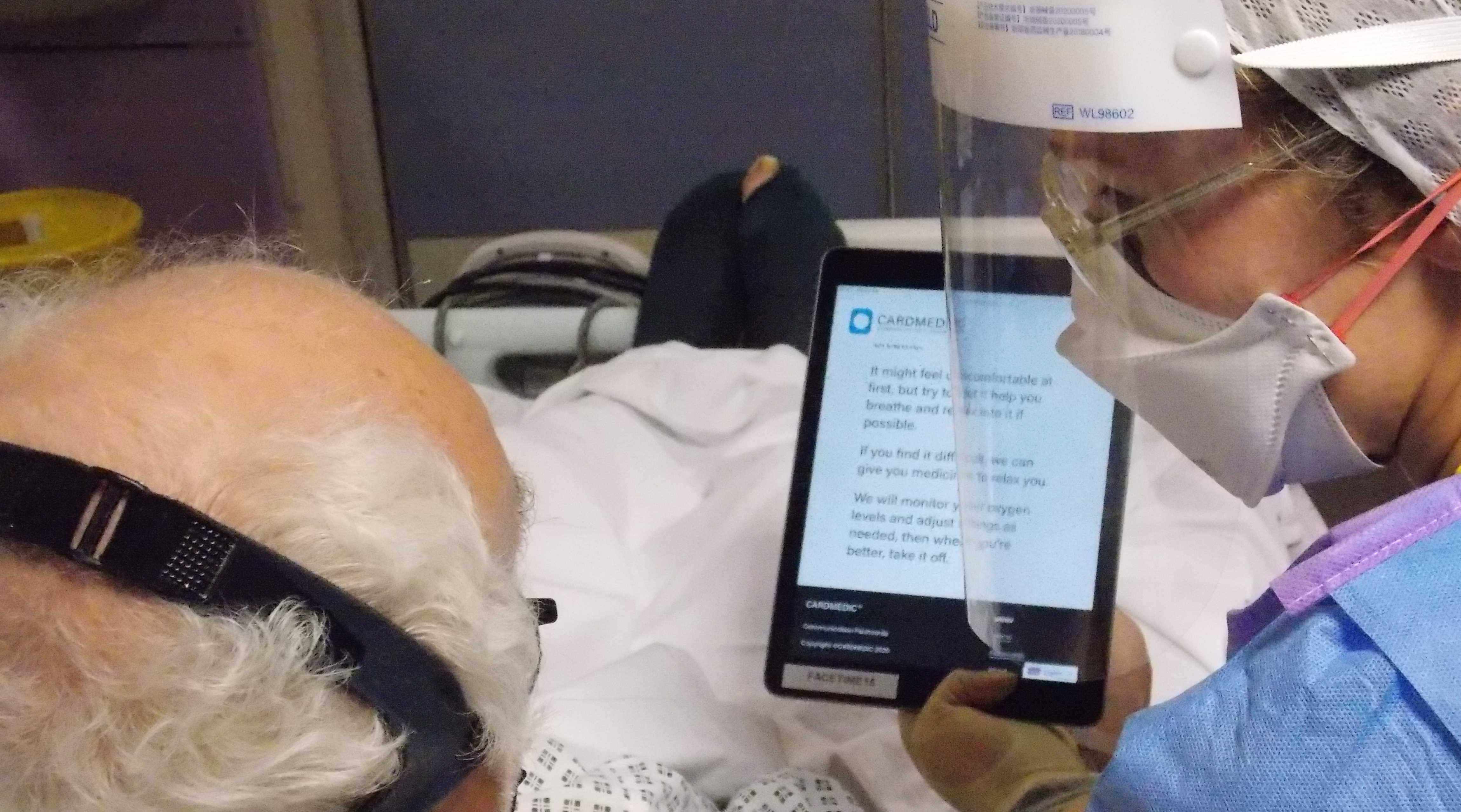 View from behind elderly patient's head of a nurse in full personal protection holding up a tablet with text explaining how oxygen treatment might make him feel. 