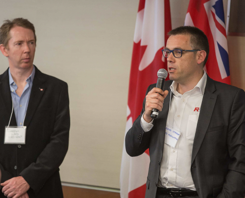 Roger Estrada, CEO of R53 Engineering, using a microphone to address an event during the GBIP visit to Canada. Looking on (left) is Gordon Jolly, ‎Industrial Technology Advisor - ‎National Research Council Canada. UK and Canadian flags on the wall behind.
