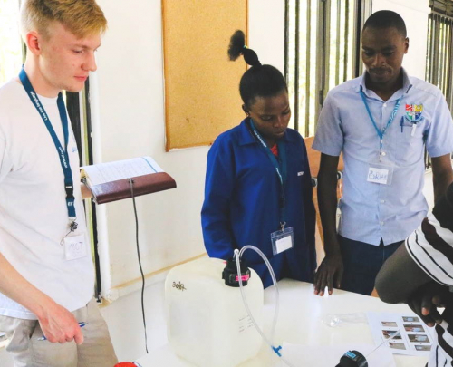 Tom Stakes, Blue Tap founder and Chief Technical Officer, holding a notepad and looking at a chlorine doser being tested in Uganda, watched by three members of the local water treatment project team, one female and two male. 